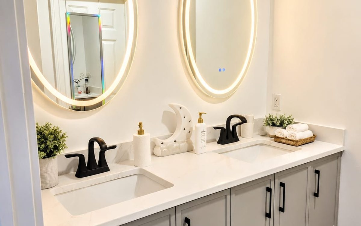 Bathroom with a grey double vanity, white quartz top, two round lit mirrors and folded towels in a basket