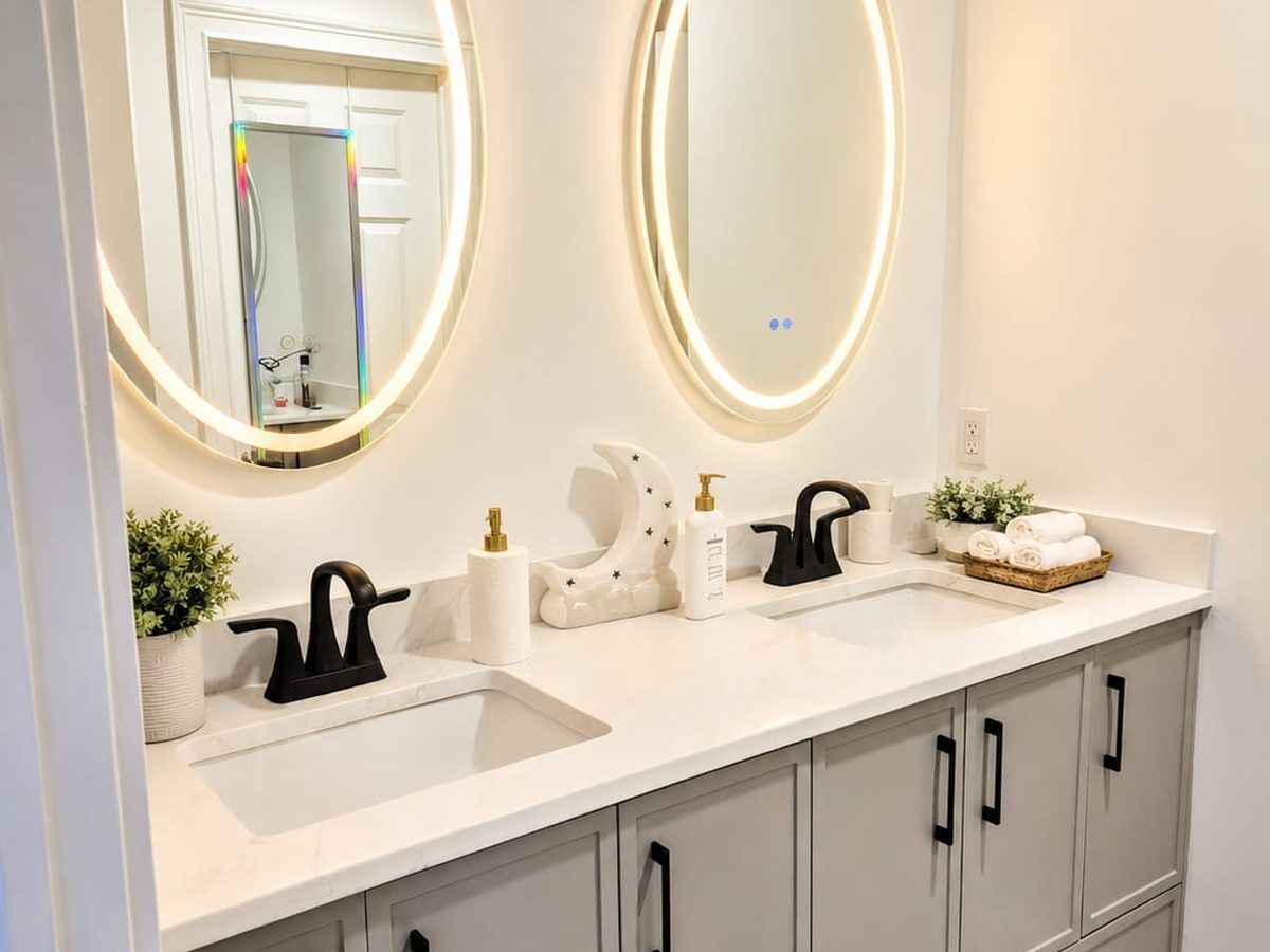 Bathroom with a grey double vanity, white quartz top, two round lit mirrors and folded towels in a basket