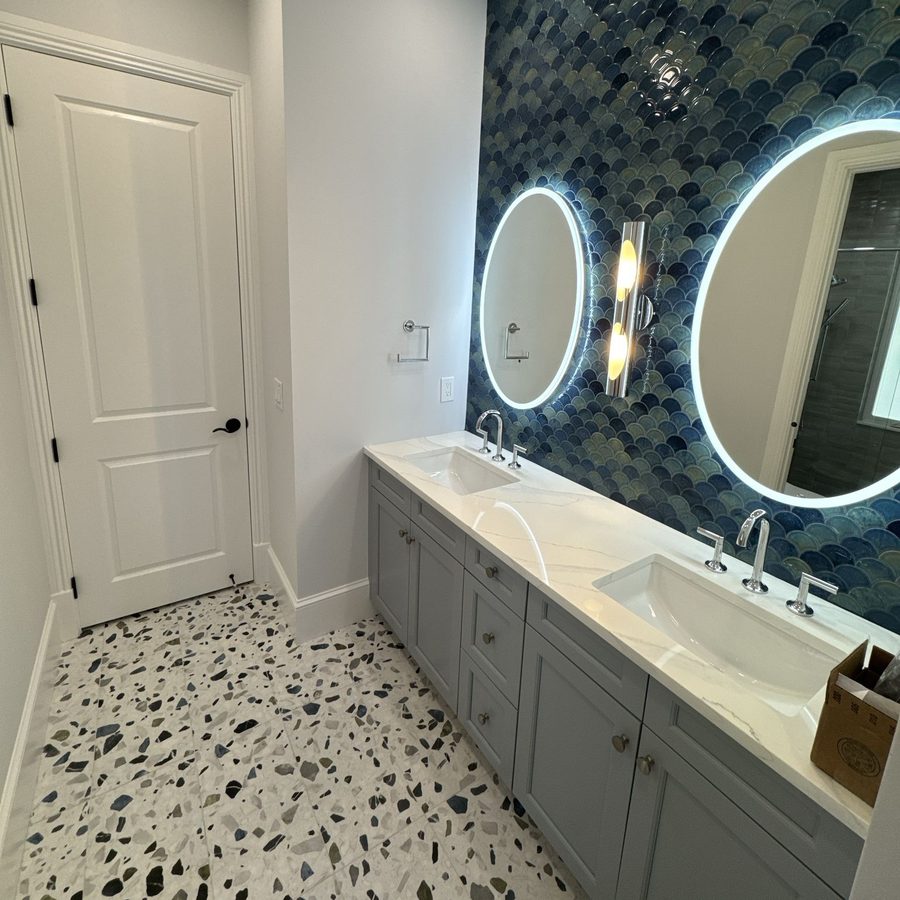 Bathroom with a blue fish-scale tile wall, two round lit mirrors, a grey double vanity and a terrazzo floor