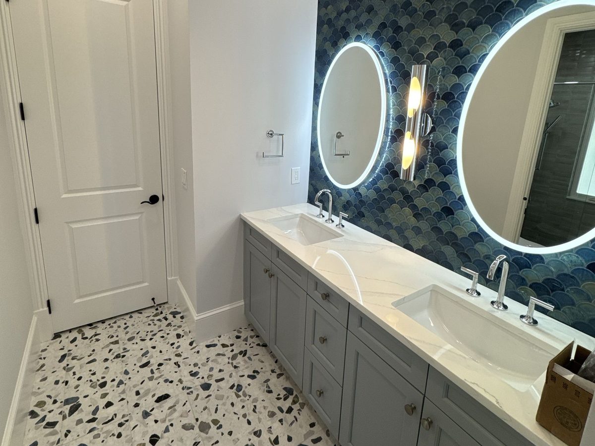 Bathroom with a blue fish-scale tile wall, two round lit mirrors, a grey double vanity and a terrazzo floor